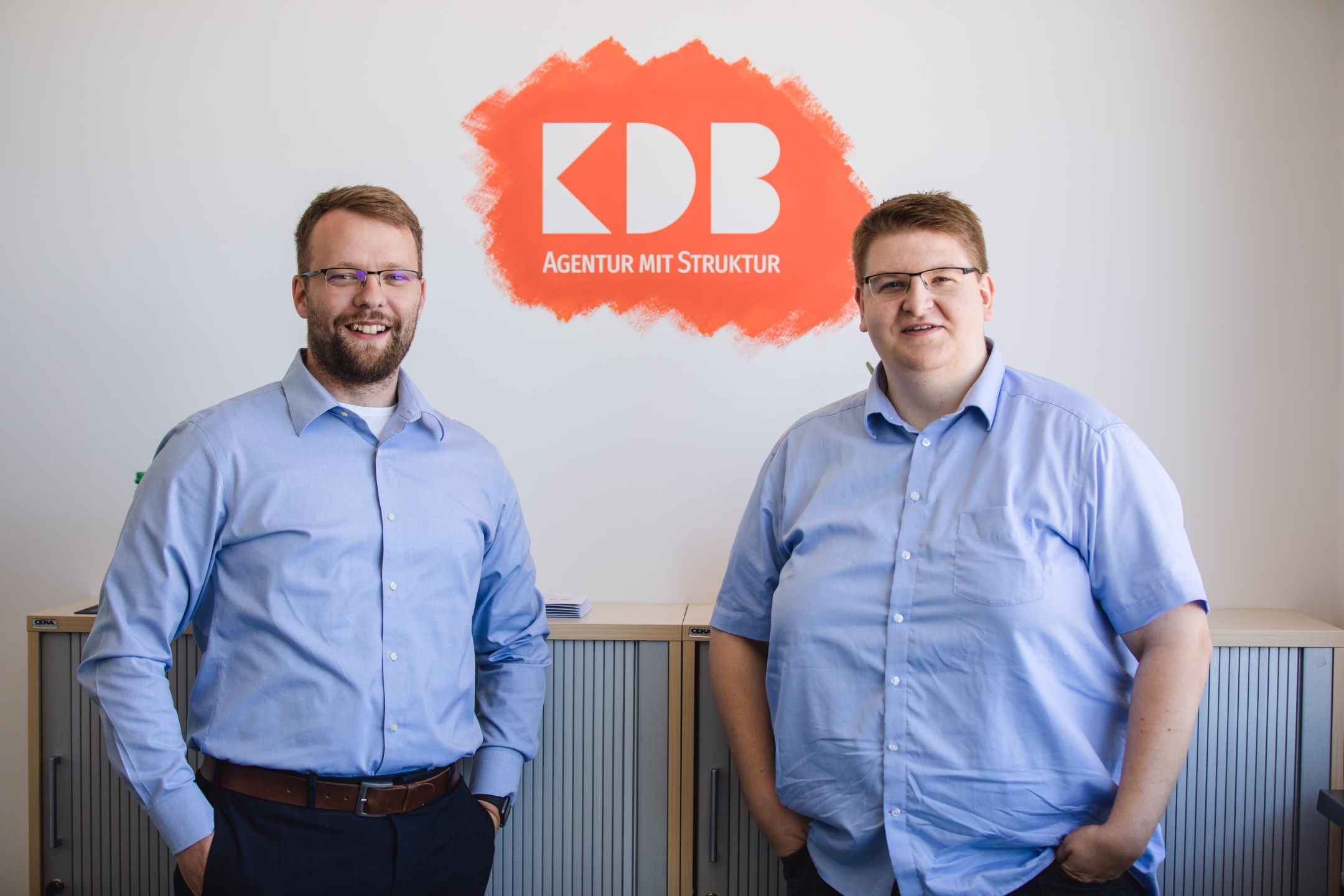 QHD_kdb_büro-12 Zwei Mitarbeiter im Büro vor orangefarbenem KDB-Logo; symbolisiert Unternehmensidentität und Teampräsenz.